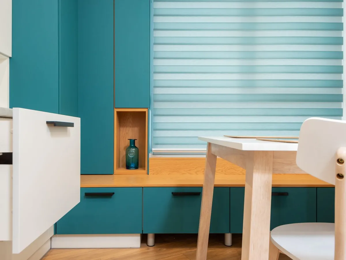 Wide light-filtering horizontal blind covering a kitchen window above teal cabinets