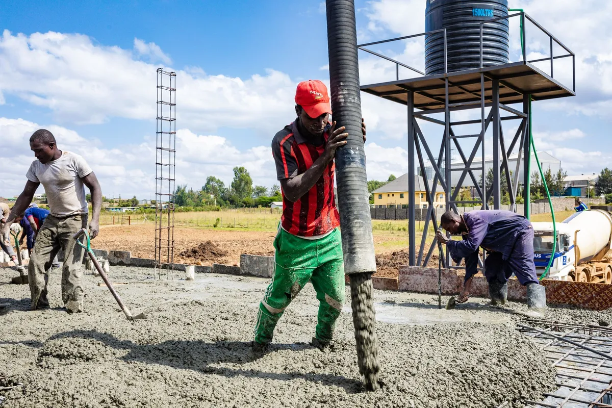 A crew placing concrete from a pump hose over a reinforced slab, with a mixer truck behind — forming, pouring and finishing labor is 35–45% of a driveway budget, and the slab is placed in a single day once the base is ready.