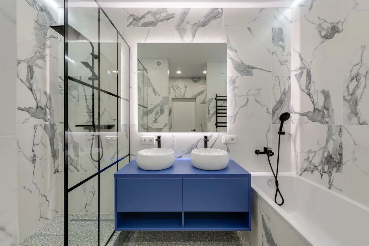A marble-look bathroom with a double vanity and a tub and shower wrapped in matching veined panels — cultured marble's project family is exactly this: molded tops and surround panels supplied as finished pieces, which is why its labor share runs below slab stone. Captions describe the visible surfaces only; the pictured bathroom is not certified as cultured marble.
