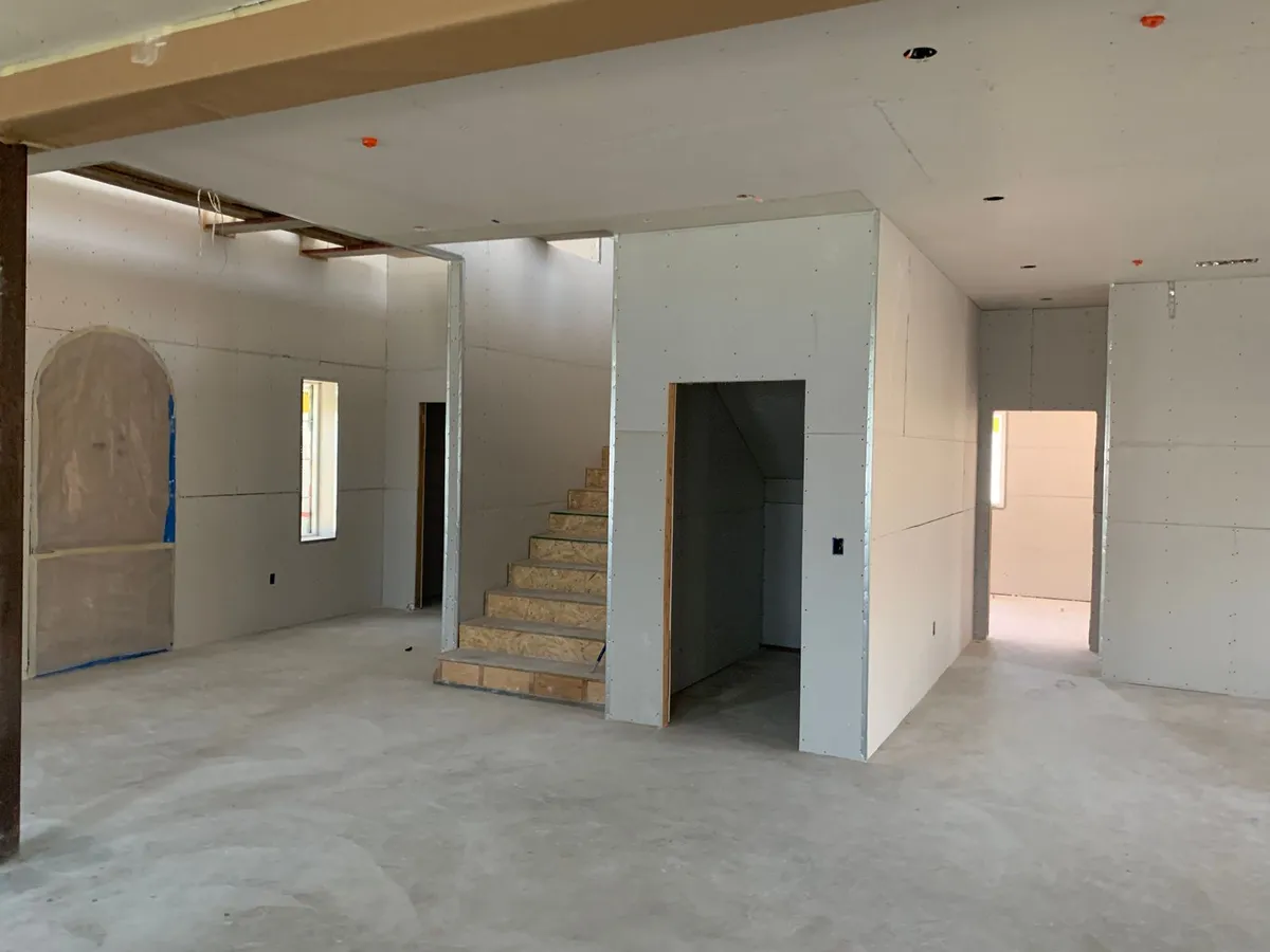 Interior under construction with freshly hung drywall walls and ceiling, panel seams and screws visible, a staircase in the centre