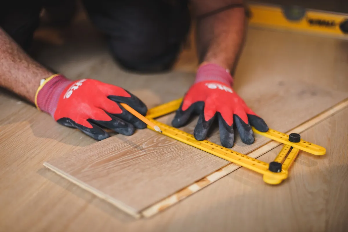 An installer measuring and marking a wood plank before cutting it — installation labor is 30–40% of an engineered hardwood budget, and accurate measuring and cutting is the part of that labor that decides how tight the finished floor looks.