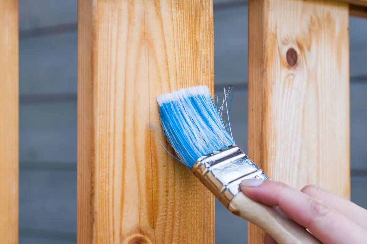A hand brush-painting an exterior wooden fence post — detail and trim work like this is slow, brush-by-brush labor, which is why labor is 65–80% of an exterior painting budget.