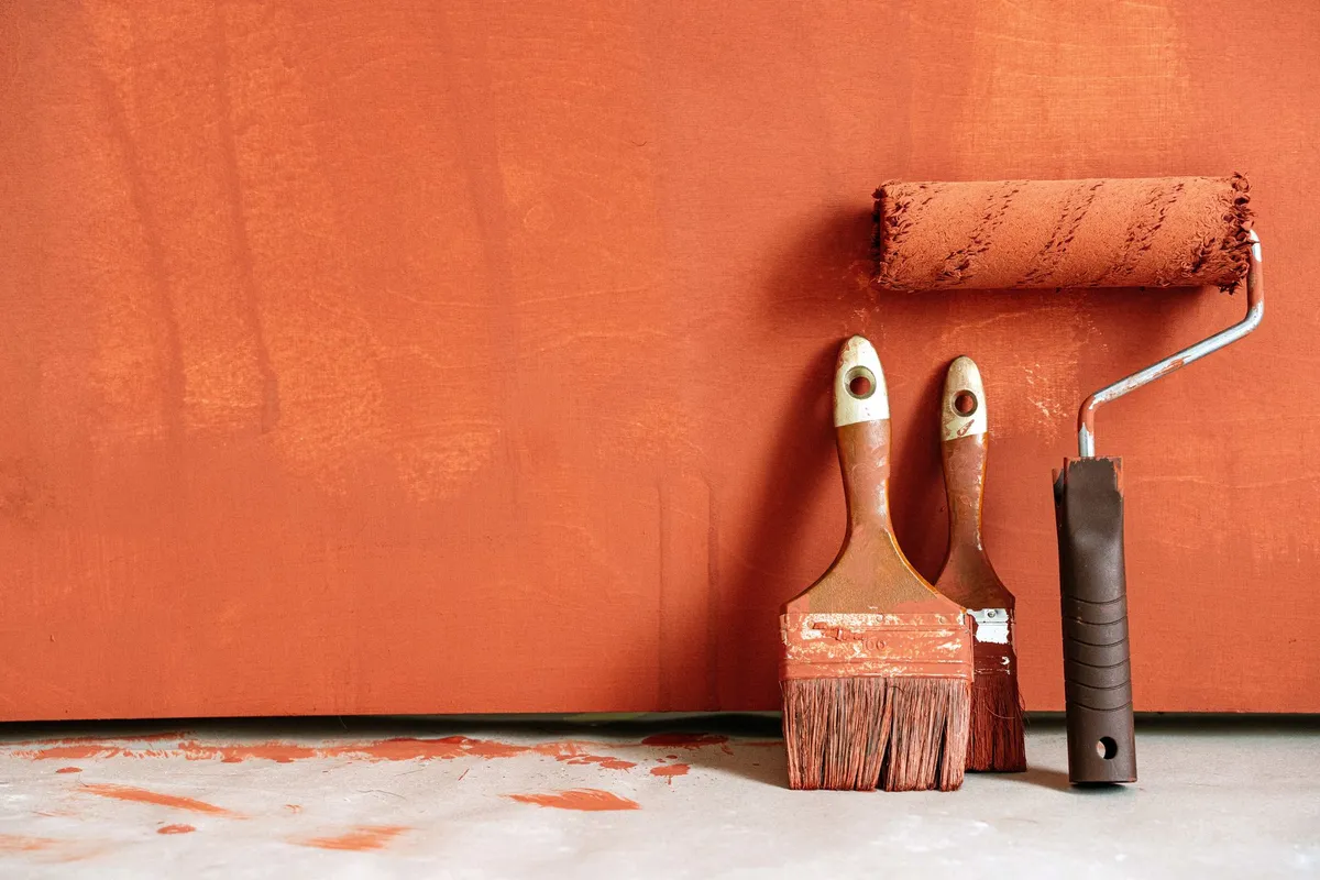 Paint brushes and a roller resting against a freshly painted terracotta wall — brushes for cutting in and a roller for the field are the standard kit; the paint itself is only 15–25% of a professional interior budget.