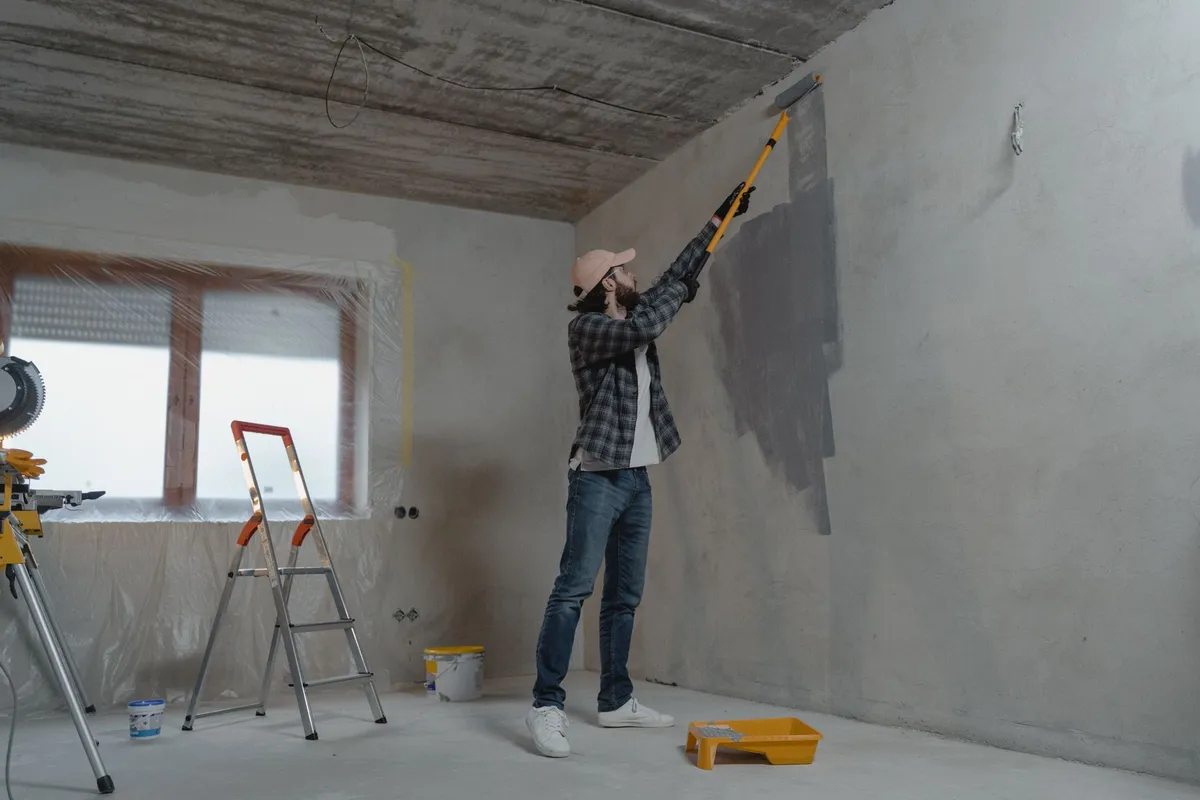 A painter rolling gray paint onto an interior wall, windows masked with plastic behind him — masking, prep and two even coats are the labor that makes up 65–80% of an interior painting budget.