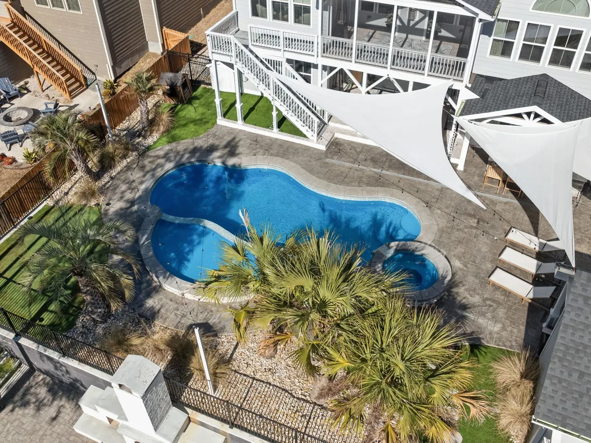 Aerial view of a freeform inground pool with an attached round spa, stamped concrete deck and shade sails