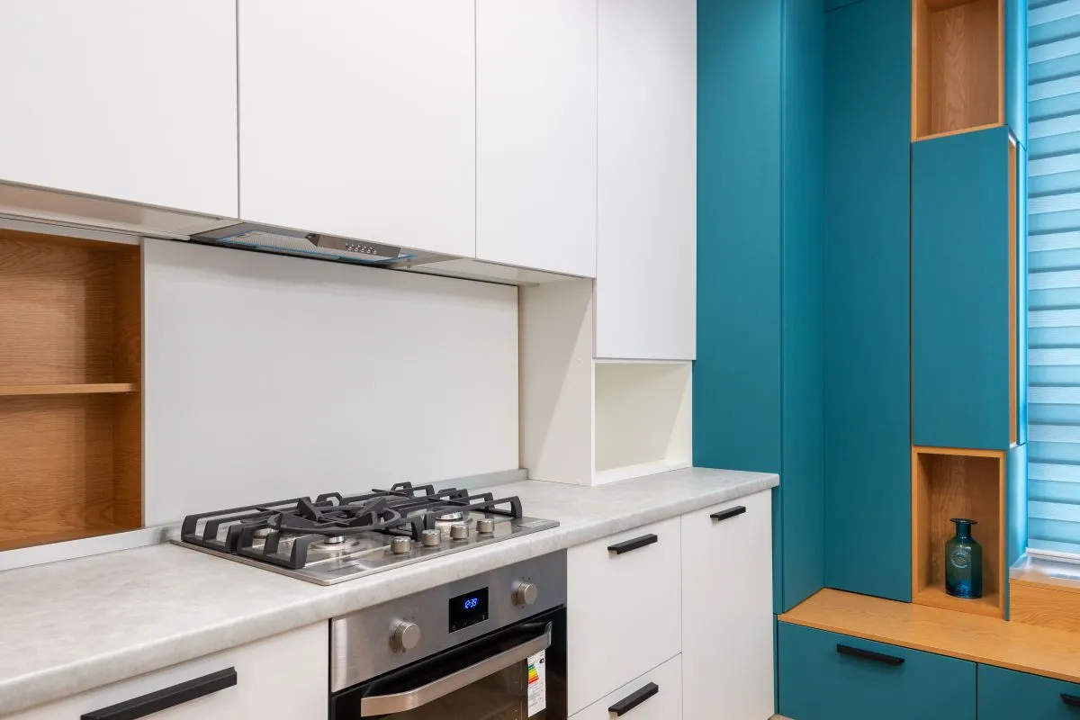 A light counter run with a gas cooktop cut into it, teal cabinets alongside — fabrication and installation labor is 35–50% of a solid-surface budget, and the seamless joins are what that skilled work buys.