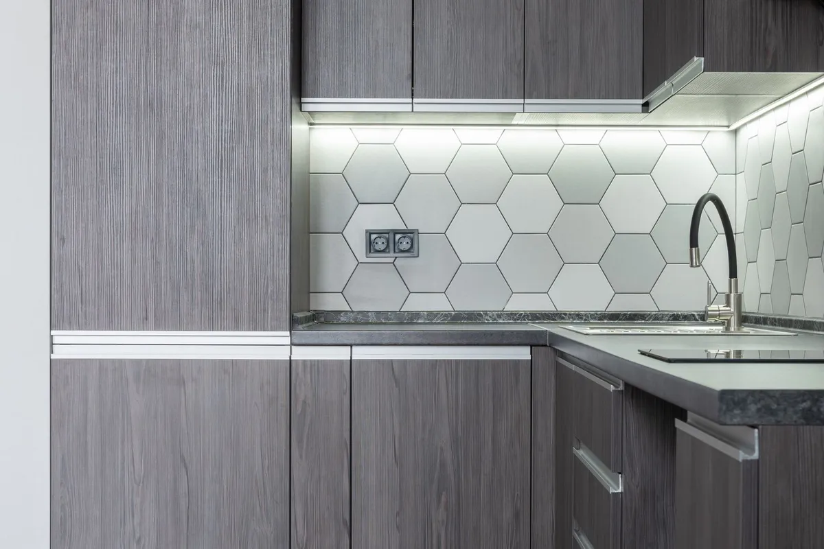 Matte dark-grey counters under grey wood-grain cabinets with a hexagonal tile splashback — zinc's appeal is exactly this flat, blue-grey metal tone, and at $90–$200 per sq ft installed the counter is the deliberate spend in an otherwise standard kitchen. The pictured counters are not certified as zinc.